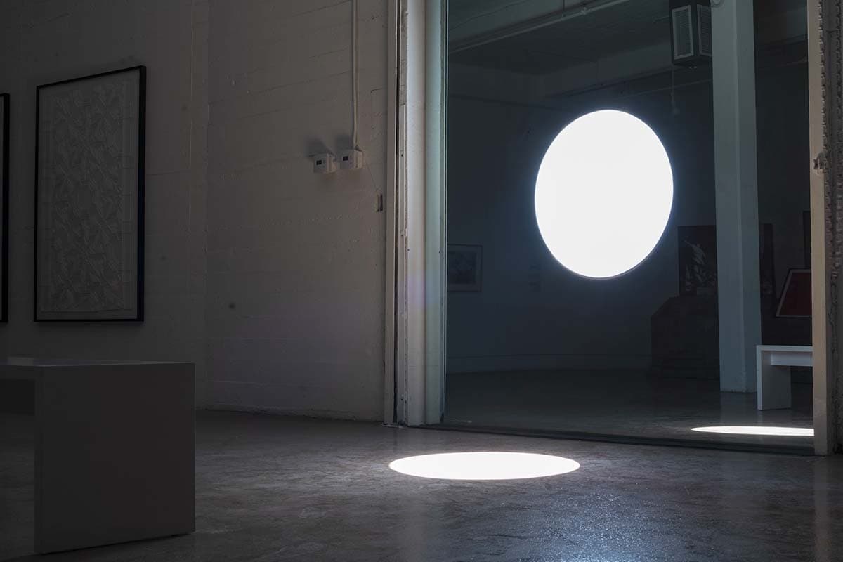 An interior space with a mirror on a wall. The mirror has a hole in the middle through which sunlight shines onto the ground.